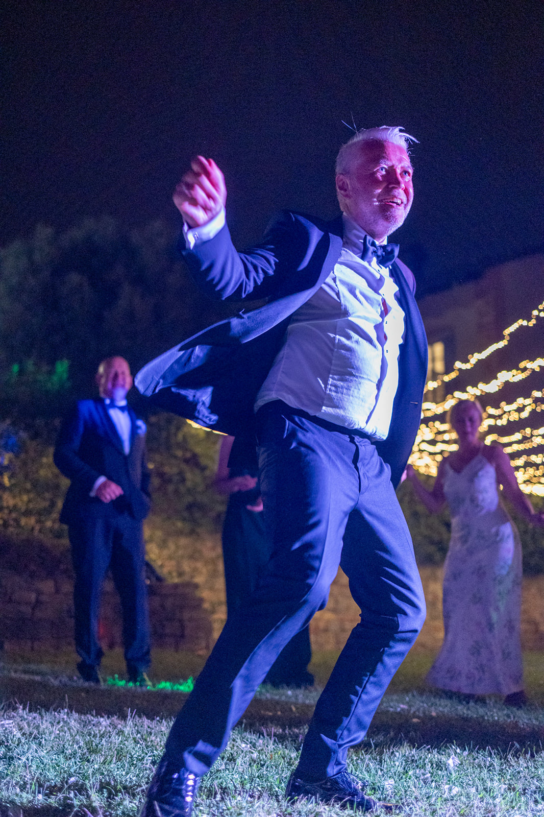 Groom in tuxedo dancing joyfully at outdoor evening wedding reception with fairy lights and guests
Groom in tuxedo dancing joyfully at outdoor evening wedding reception with fairy lights and guests