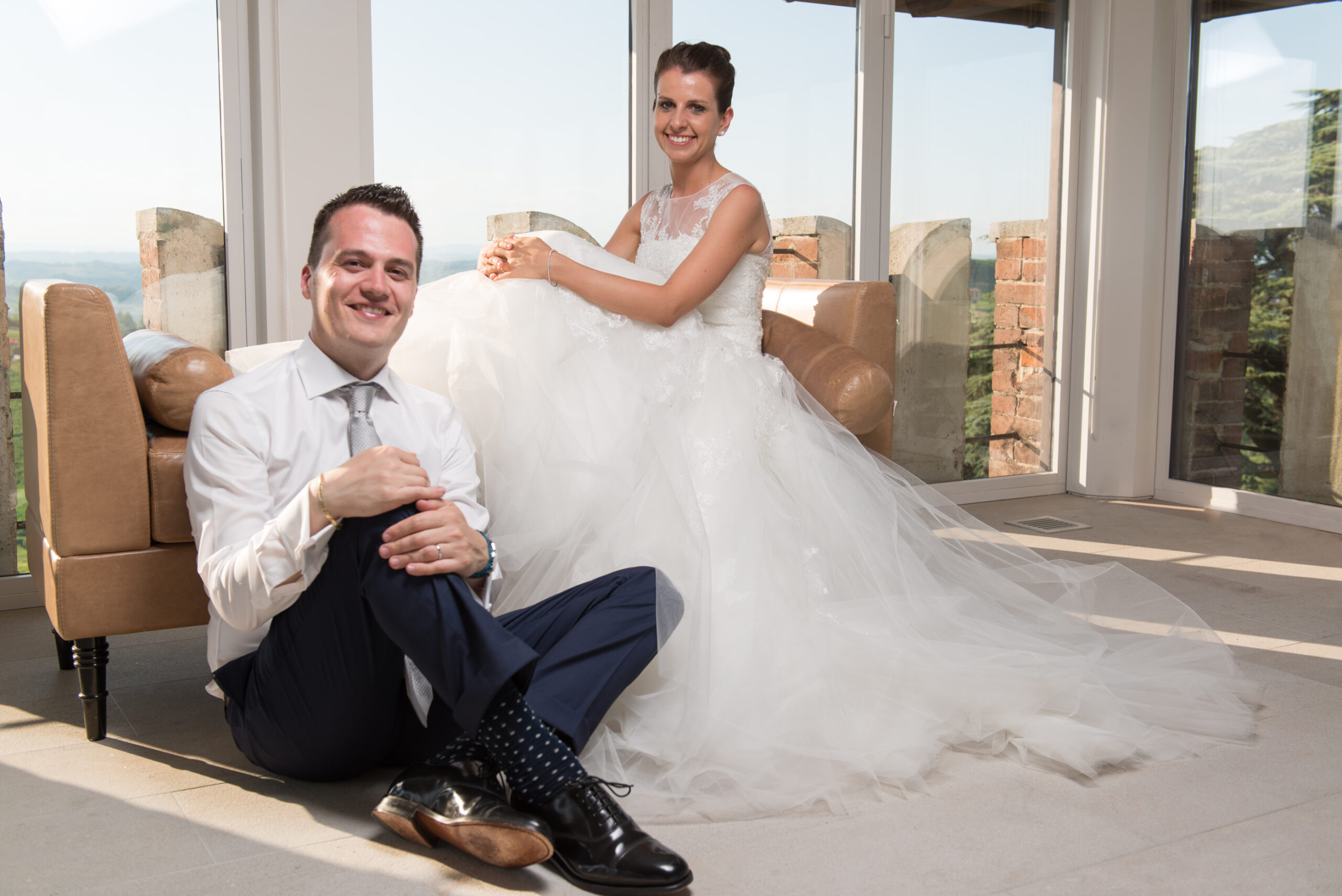 Smiling bride and groom relaxed portrait by sunlit windows overlooking Italian countryside vineyard hills